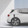 White electric vehicle connected to a tall white charging station with a cable plugged into the rear side of the car.